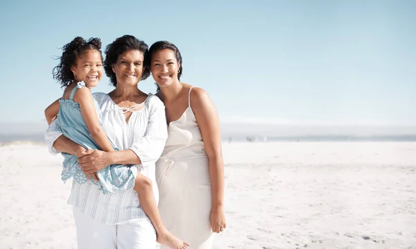Smiling mixed race family standing together on a beach with copyspace. Happy hispanic grandmother bonding with granddaughter over weekend. Adorable little girl enjoying free time with single mother.