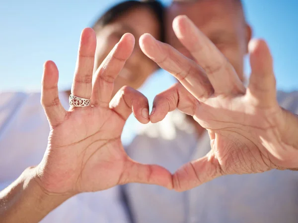 Affectionate mature mixed race couple sharing an intimate moment on the beach. Senior husband and wife making a heart shape with their hands. They love spending time together by the sea at sunset.