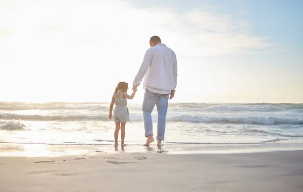 Rearview cute mixed race girl standing hand in hand with her father in the sea at the beach. A young man and his daughter holding hands while standing in the water and looking at the view at sunset.