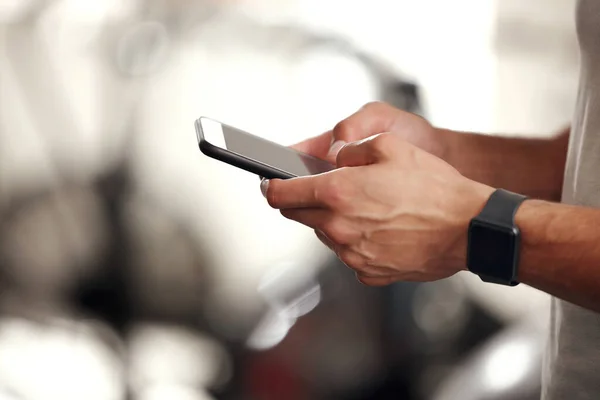 Closeup of one fit mixed race man using a cellphone while taking a break from exercise in a gym. Hands of a guy texting and browsing fitness apps online while checking social media during a rest from