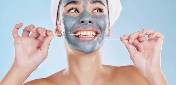 Beautiful young mixed race woman flossing her teeth with dental floss for oral hygiene isolated in studio against a blue background. Fresh and clean female out the shower, wearing a face mask peel.