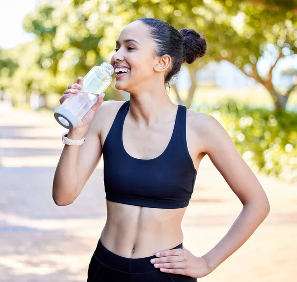 One fit young mixed race woman taking a rest break to drink water from bottle while exercising outdoors. Happy female athlete quenching thirst and cooling down after running and training workout.