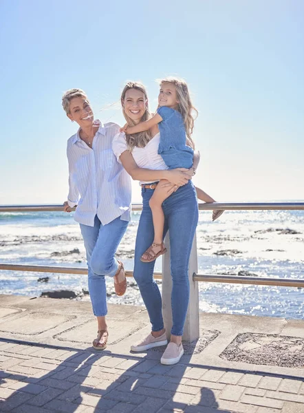 Full length of female family members posing together at the beach on a sunny day. Grandmother, mother and granddaughter standing together on seaside promenade. Multi-generation family of women and