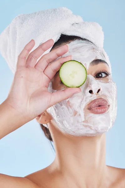 Beautiful young mixed race woman wearing a face mask peel and towel while holding cucumber slices. Attractive female clean and fresh out the shower and applying her daily skincare regime to her face.