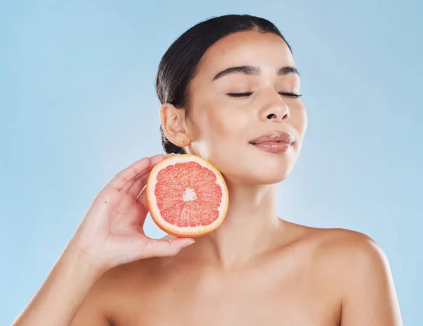 Beautiful young mixed race woman with a grapefruit isolated in studio against a blue background. Her skincare regime keeps her fresh. For glowing skin, eat healthy. Packed with vitamins and nutrients.