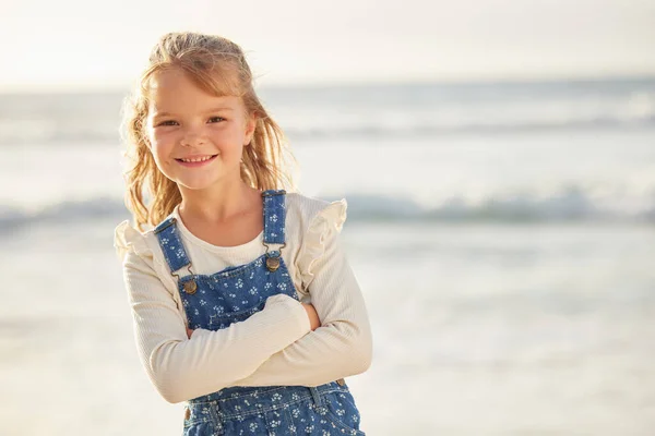 Happy little girl standing with her arms crossed during summer at the beach. Cheerful caucasian small child enjoying a day at the beach, posing. Blond girl enjoying a sunny summer day by the ocean.