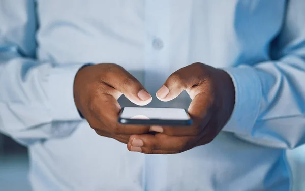 Closeup hands of african man using a phone while standing in his office. African american business man sending a text message while working late at night at his work. Putting in overtime after hours.