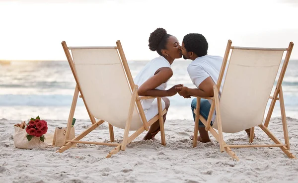 Happy african American couple spending a day at the sea together. Content boyfriend and girlfriend kissing while sitting on the beach. Caring husband and wife bonding on the seashore.