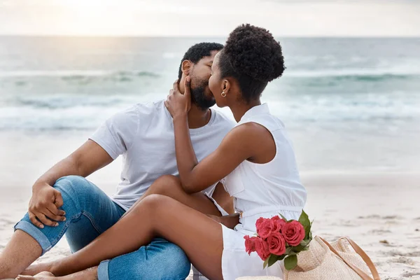 African American couple spending a day at the sea together. Content boyfriend and girlfriend kiss while sitting on the beach. Caring husband and wife bonding on the seashore.