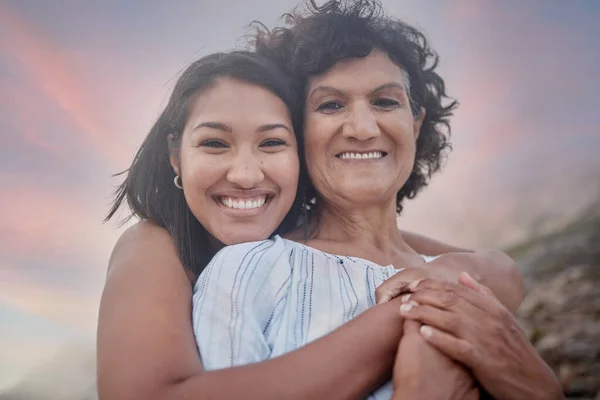 Portrait of a young hispanic woman spending the day at the beach with her elderly mother. Mixed race female and her mother smiling at the beach and hugging each other.