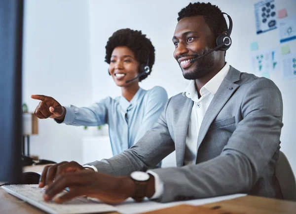 Happy african american male call centre telemarketing agent discussing plans with colleague while working together on computer in an office. Two consultants troubleshooting solution for customer