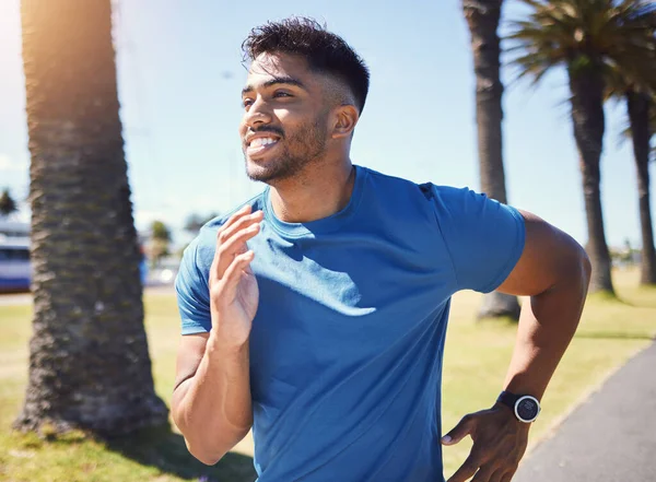 Happy smiling young mixed race fit man enjoying a run in a park during summer alone. One cheerful hispanic male smiling, exercising and jogging in nature. Dedicated to fitness and a healthy lifestyle.