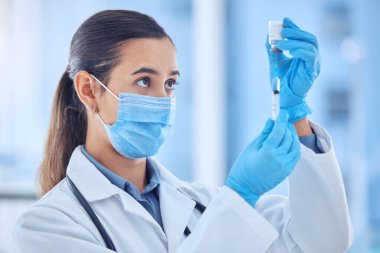 Genç melez kadın doktor hastanede aşı hazırlarken maske takıyor. Ciddi bir İspanyol kadın doktor ayakta duruyor ve klinikteki şırıngayla bir şişeden sıvı çıkarıyor..
