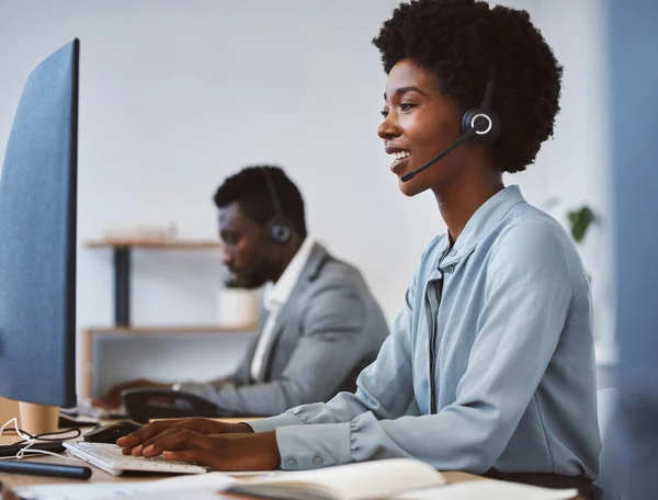 Happy young african american call centre telemarketing agent talking on a headset while working on computer in an office. Confident friendly female consultant operating helpdesk for customer service