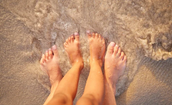 Above shot of the feet of a caucasian couple in the ocean while standing on the sand at the beach. Romantic people sharing an intimate moment during a date by the seaside. Love and togetherness.