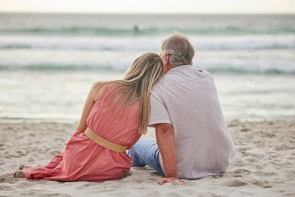 Rear view of a caucasian father and daughter sitting on the sand at the beach together and bonding looking at the view.