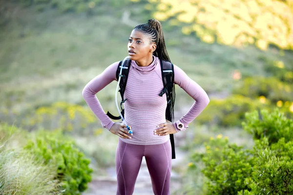 Young fit and active african american woman hiking through the mountains and catching her breath. Black woman standing alone with her hands on her hips and carrying a backpack while exploring nature.