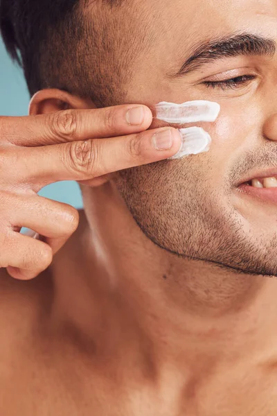 Closeup of one young indian man applying moisturiser lotion to his face while grooming against a blue studio background. Handsome guy using sunscreen with spf for uv protection. Rubbing facial cream