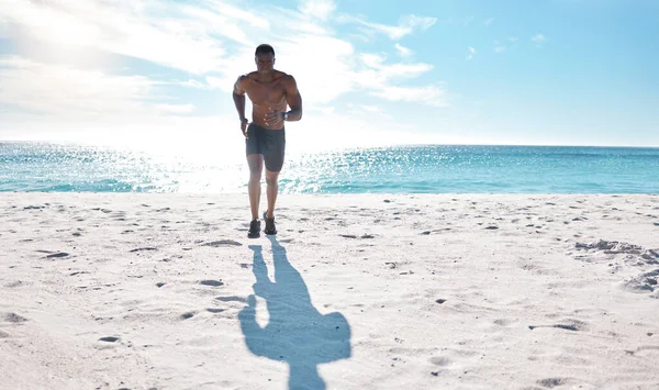 Fit young black man running and jogging on sand at the beach in the morning while exercising. One strong male bodybuilder athlete with six pack abs doing cardio workout to build muscle and endurance.