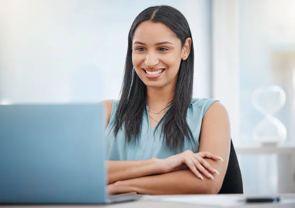 Happy young mixed race businesswoman smiling while enjoying working on a laptop sitting in a chair in an office at work. One hispanic female boss sending an email using a laptop at a desk at work