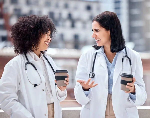Two happy and cheerful female doctors standing and drinking a cup of coffee outside taking a break together. Caucasian and mixed race hispanic women smiling relaxing and talking on a break outside.