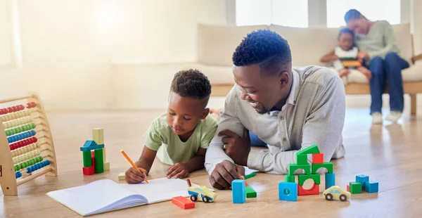 Young happy african american father helping his son with homework while sitting on the floor at home. Little boy focused on writing in a notebook and doing a task. Small boy drawing in a book.