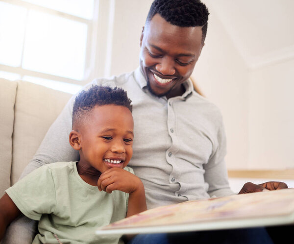 African american father bonding with his at home. Black male helping his son read a book and practice learning while sitting on a sofa at home. cute little black boy reading to his dad in the lounge