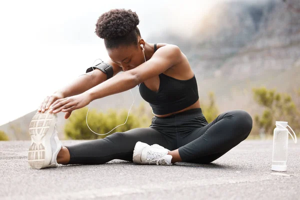 One african american female athlete with an afro listening to music on her earphones while exercising outdoors in nature. Dedicated black woman smiling while warming up before a workout outside.