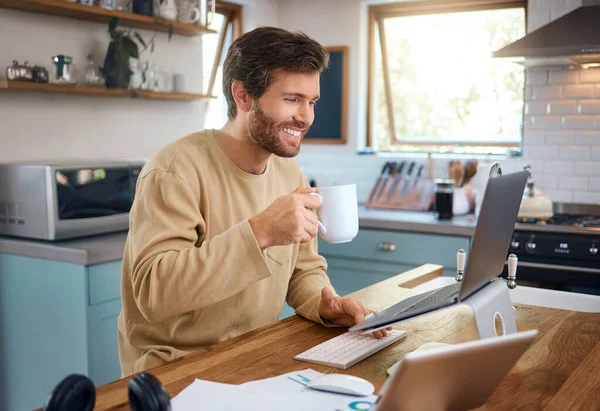 Happy young caucasian man drinking coffee while working on laptop in kitchen, checking his email or searching information while doing freelance work at home. Smiling young male using internet banking