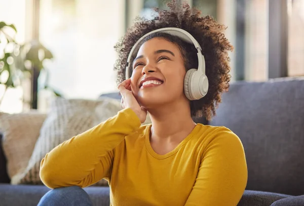 Young cheerful mixed race woman thinking while wearing headphones and listening to music at home. One content hispanic female with a curly afro enjoying music and daydreaming while sitting on the