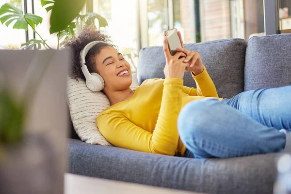 Young joyful mixed race woman wearing headphones and listening to music while typing a message on a phone at home. One content hispanic female with a curly afro using social media on a cellphone while