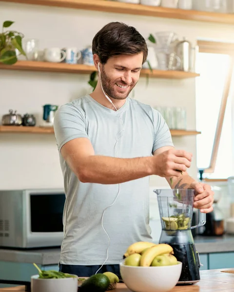 One fit young caucasian man adding ingredients to blender to make healthy green detox smoothie while wearing earphones in kitchen at home. Guy having fresh fruit juice to cleanse and provide energy