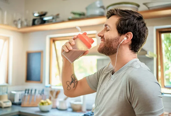 One fit young caucasian man drinking bottle of chocolate whey protein shake for energy for training workout while wearing earphones in a kitchen at home. Guy having nutritional sports supplement for