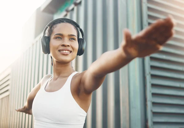 One fit young hispanic woman stretching arms in warrior pose for warmup to prevent injury while exercising in an urban setting outdoors. Happy and motivated female athlete listening to music with