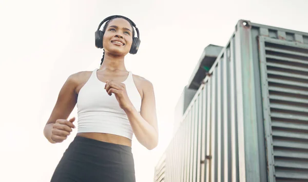 One active young hispanic woman from below listening to music with headphones while running in an urban setting outdoors. Happy female athlete doing cardio workout while exercising for better health