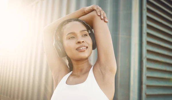 One fit young hispanic woman stretching arms for warmup to prevent injury while exercising in an urban setting outdoors. Happy and motivated female athlete listening to music with headphones while