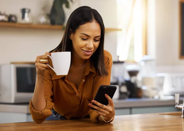 Young mixed race woman drinking a cup of coffee while using her phone alone in the morning in the kitchen. Content hispanic female smiling and enjoying a cup of tea while using social media on her