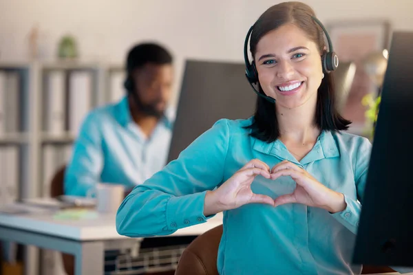 Portrait of one happy young smiling caucasian call centre telemarketing agent gesturing heart shape with her hands while talking on headset in office. Confident friendly businesswoman operating