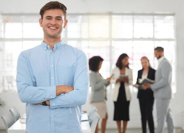 Young happy caucasian businessman standing with his arms crossed while in an office with colleagues. Content male boss In a meeting with coworkers.