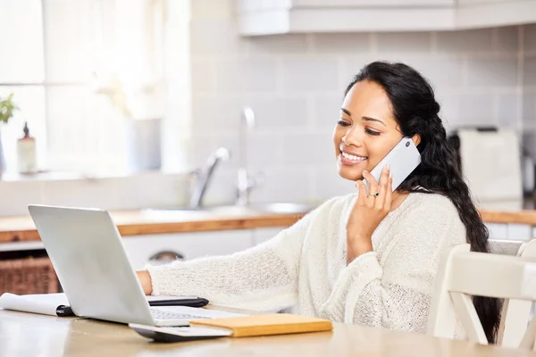 One mixed race woman working remotely using a laptop and talking on a cellphone while writing and making notes in a notepad, sitting at a table at home. Positive freelance worker doing research.