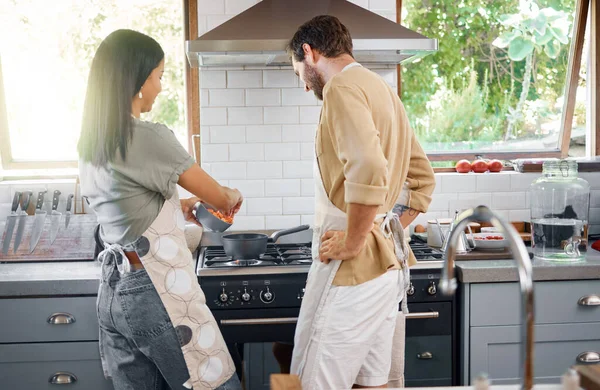 Rear view of young interracial couple cooking a healthy meal together at home. Happy young hispanic wife teaching her husband to cook as throwing vegetables into a pot on the stove.