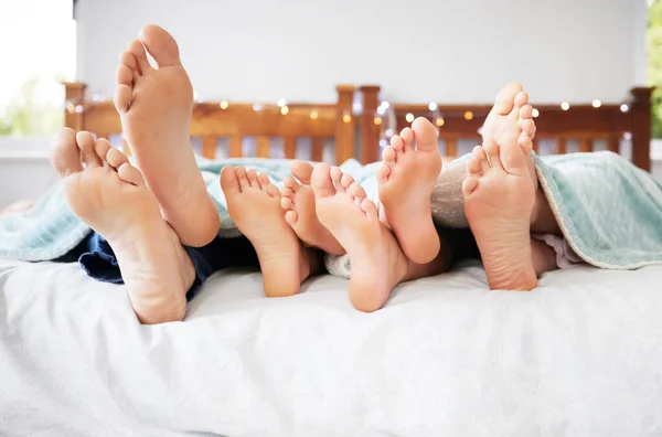 Feet of family lying in bed. Closeup of feet of parents and children in bed. Family relaxing in bed together. Below bare feet of family in bed. Kids resting in bed with their parents.