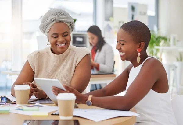 Smiling african american business women sitting together and using a digital tablet during a brainstorm meeting in the office. Confident happy black professionals planning a strategy with technology.