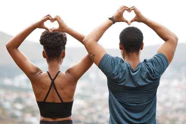 Rearview fit african american couple making heart shapes with their hands while exercising outdoors. Young athletic man and woman promoting health and fitness outside. They love to workout together.