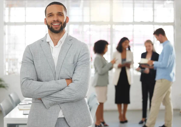 Young happy mixed race businessman standing with his arms crossed while in an office with colleagues. Hispanic male boss In a meeting with coworkers.
