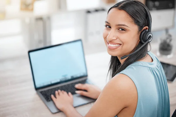 Portrait of a young mixed race female wearing a headset while working on a computer in her kitchen at home. Young hispanic female call center agent helping a customer while working at a desk.