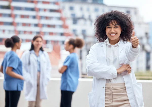 Portrait of a happy young mixed race doctor gesturing thumbs up with her colleagues in the background. Group of doctors and nurses collaborating in meeting. African american woman giving approval.