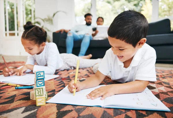Little boy and girl drawing with colouring pencils lying on living room floor with their parents relaxing on couch. Little children sister and brother siblings colouring in during family time at home.