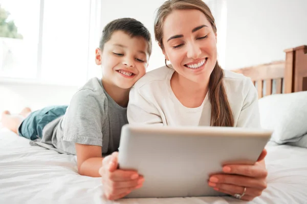 Happy caucasian mother and son holding digital tablet while lying together on a bed. Little boy and mom watching movie online or playing game while spending time at home.