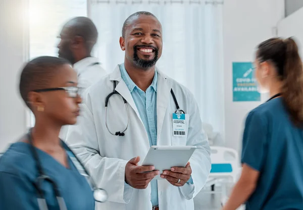 Portrait of a happy mature african american male doctor working on a digital tablet while working at a hospital with colleagues. Medical professional doing research on a digital tablet at work at a